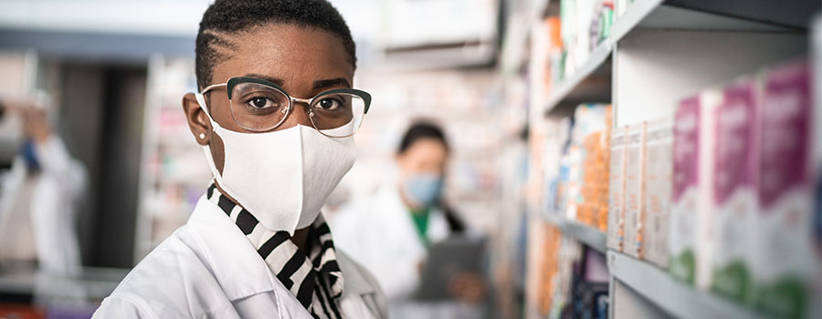 african american young woman wearing a mask and lab coat in an aisle of a pharmacy with background blurred out